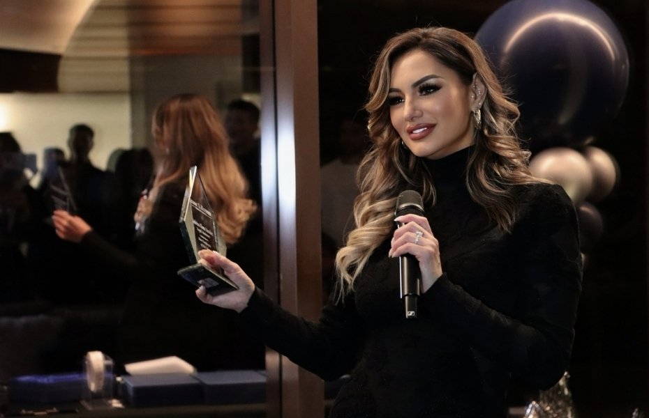 Venessa Patino speaks into a microphone while holding an award plaque, with holiday balloons in the background.