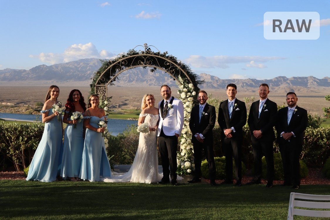 Bride in lace gown and groom in white tuxedo stand beneath a floral arch with their bridesmaids in blue dresses and groomsmen in black suits, overlooking the desert mountains of Paiute Golf Course.