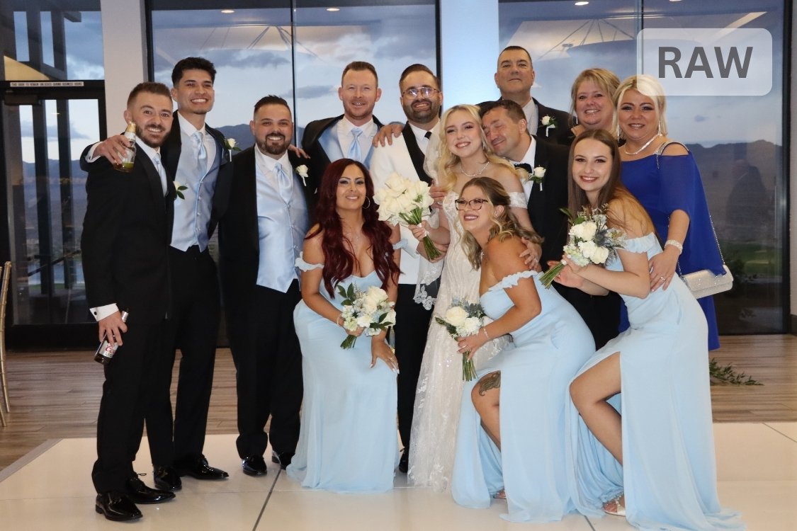 Indoor group photo of the full wedding party: bride in lace gown and groom in white tuxedo surrounded by smiling bridesmaids in pale blue dresses and groomsmen in dark suits.