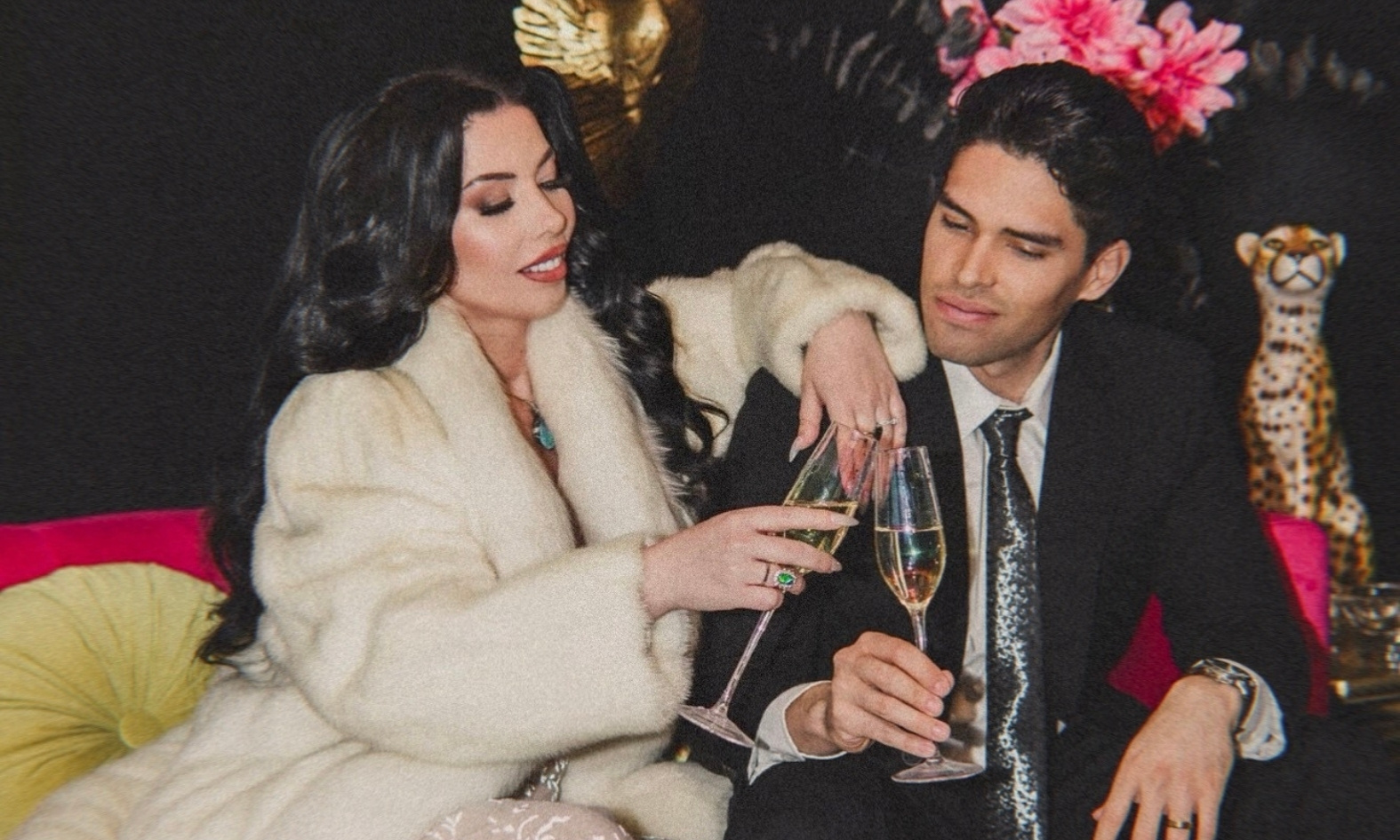 Royilda Medina and Marlon Medina sit together on a vibrant pink sofa, dressed in formal attire, raising champagne glasses in a celebratory toast within a luxurious, art-deco styled room.