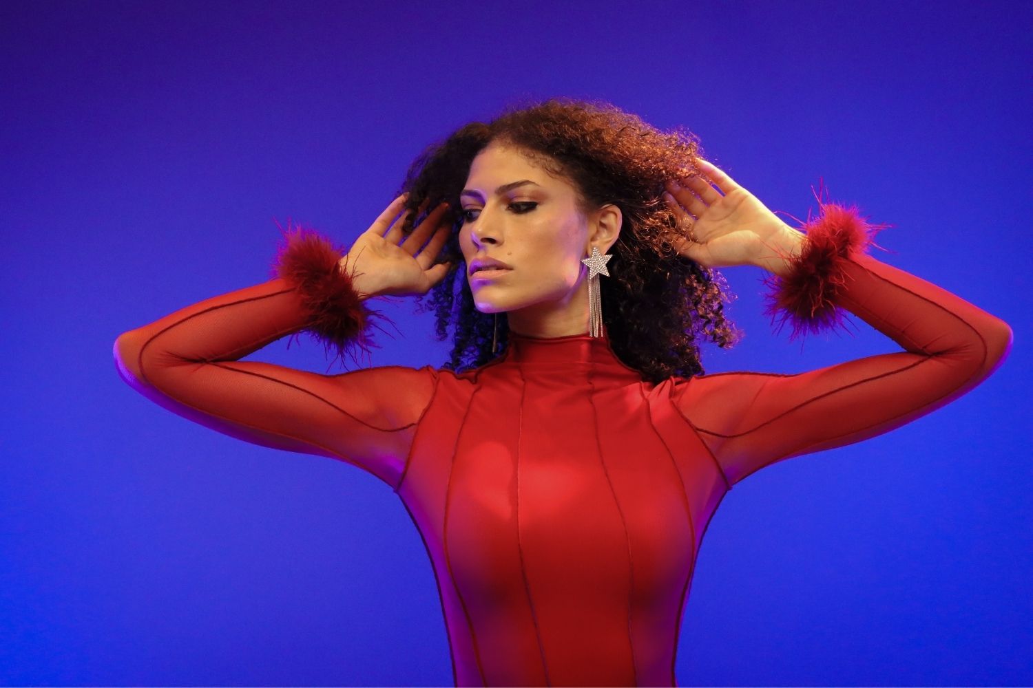 Woman in red fashion dress posing with arms raised against a blue-purple studio background, styled for an editorial portrait