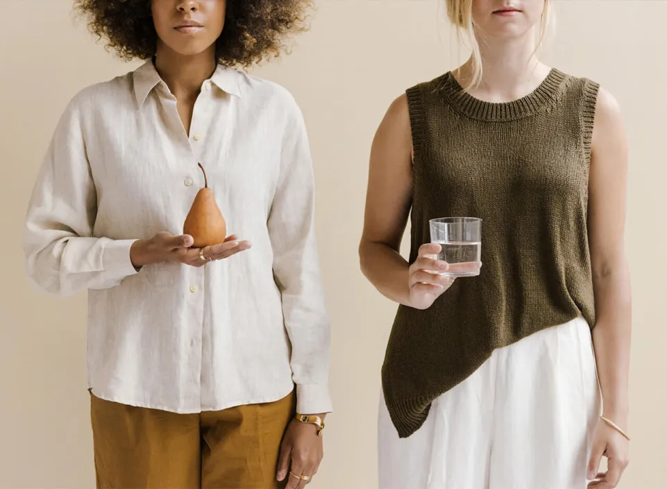 Two women standing, one holding a pear and the other holding a glass of water, against a neutral background.