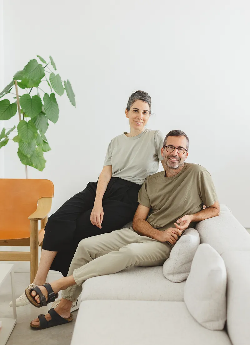 A man and a woman sitting closely together on a light gray sofa in a bright room with white walls, a wooden chair with an orange seat, and a large green plant, both smiling and casually dressed.