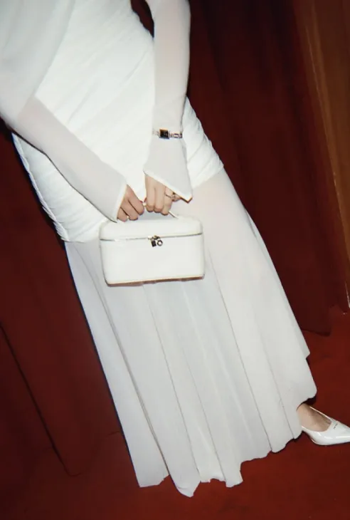 Person in white long dress holding a white handbag, standing on red carpet with maroon curtain backdrop.
