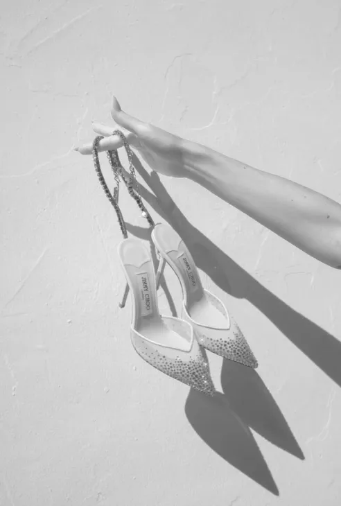 Hand holding a pair of pointed-toe high heel shoes with chains against a textured wall.