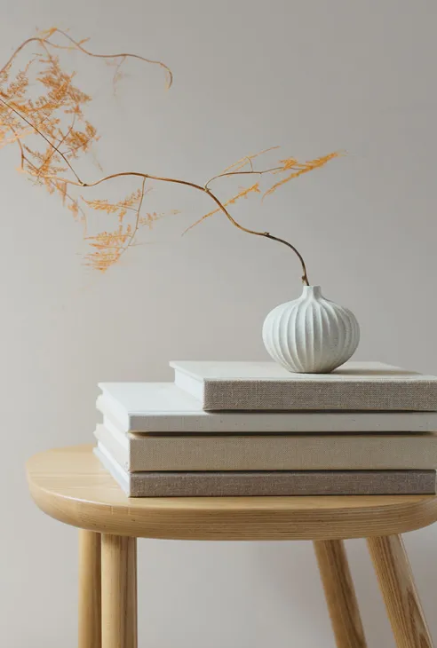 White vase with dried branch on stacked books atop a wooden round table against a plain wall.