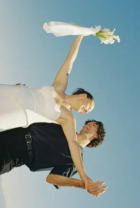Bride and groom smiling, bride holding flowers up against a clear blue sky.