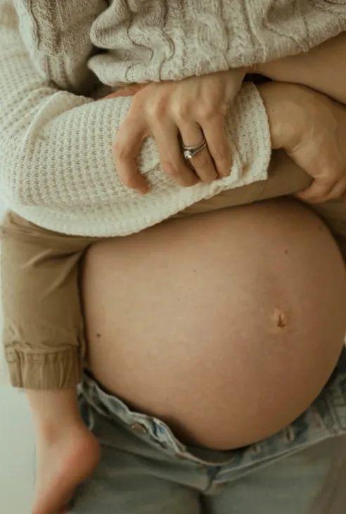 Close-up of a pregnant belly with a child’s arms wrapped around it and a hand with a ring resting on the child’s arm.