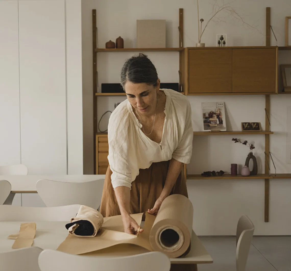 Woman in a beige blouse and brown skirt unrolling brown paper on a white table in a modern room.