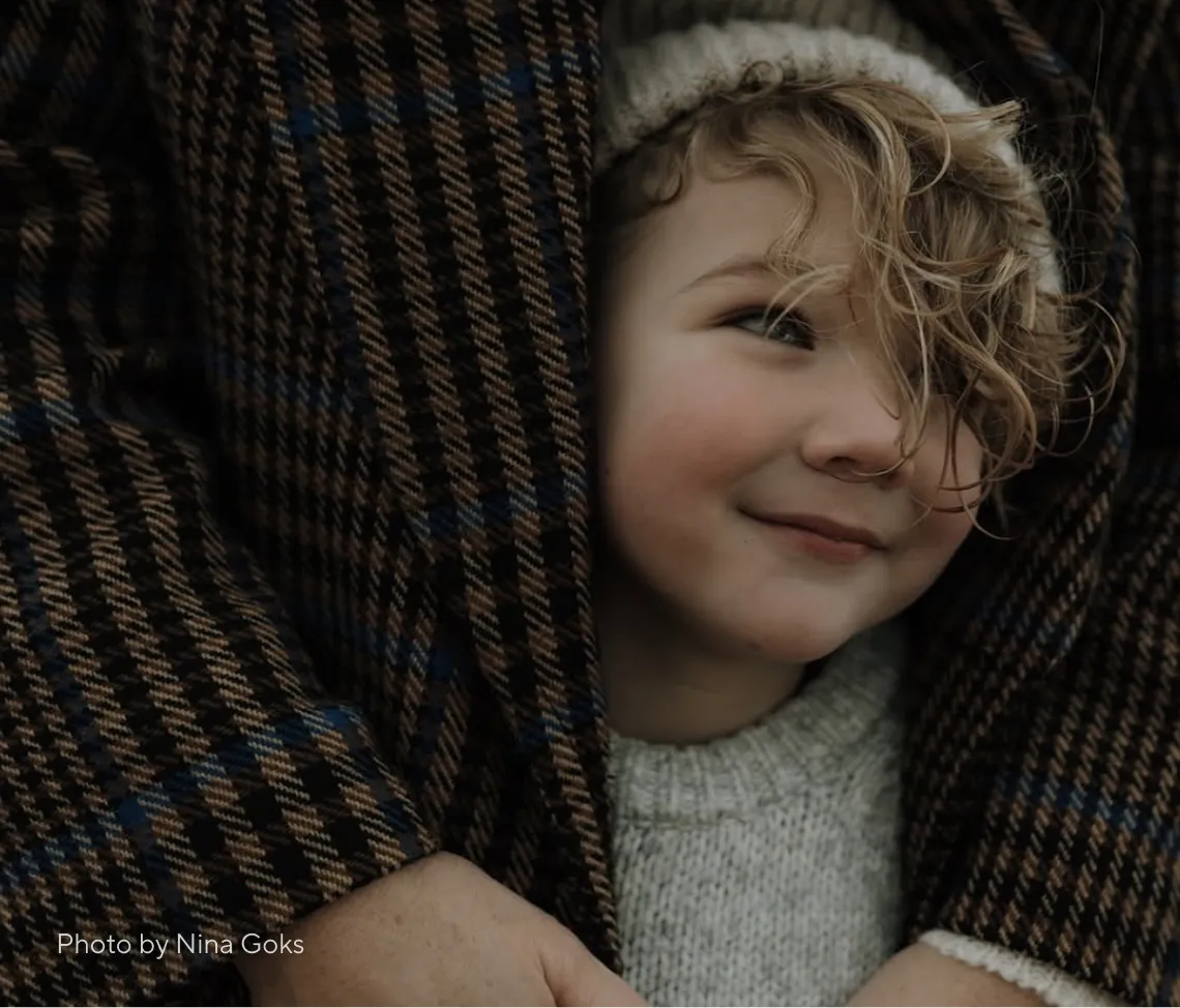 Smiling child with curly hair wearing a knit hat and sweater hugged by someone in a checkered coat.