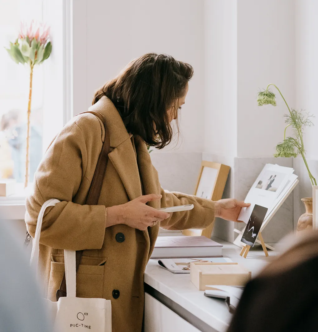 Woman in brown coat holding a phone and looking at a photo display on a white countertop.