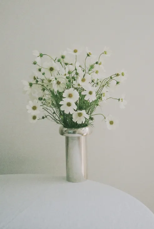 White daisies with green stems arranged in a silver vase on a white surface against a plain wall.