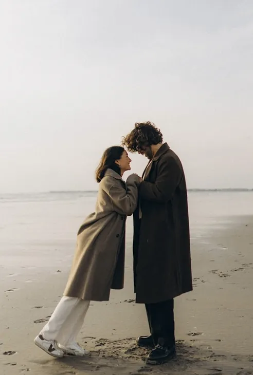 Couple wearing long coats holding hands and looking at each other on a beach.