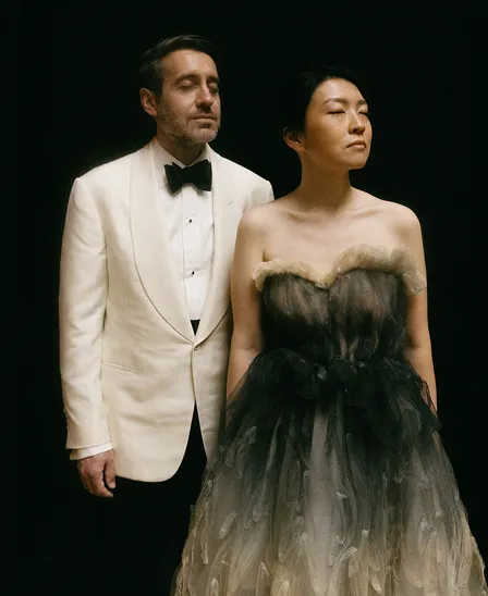 Man in white tuxedo and woman in strapless black and beige dress stand with eyes closed against black background.
