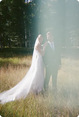 Bride in white gown and groom in suit standing together in a sunlit grassy field with trees.