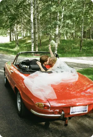Newlywed couple kissing in a red convertible car with the bride holding a bouquet up.
