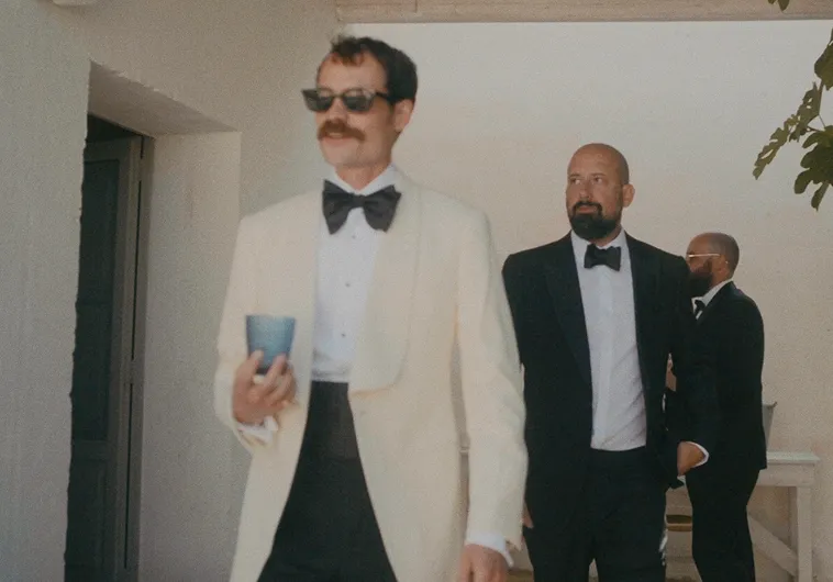 Three men in formal attire: a man in a white tuxedo jacket with sunglasses and a beard holding a glass, and two men in black tuxedos, one bald with a beard looking to the right, and another with glasses facing away, standing in a shaded outdoor area.