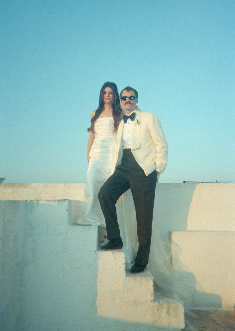 A couple dressed in formal wedding attire stands on white concrete steps against a clear blue sky; the woman wears a strapless white gown and the man, with a mustache and sunglasses, wears a white tuxedo jacket with black pants and bow tie.