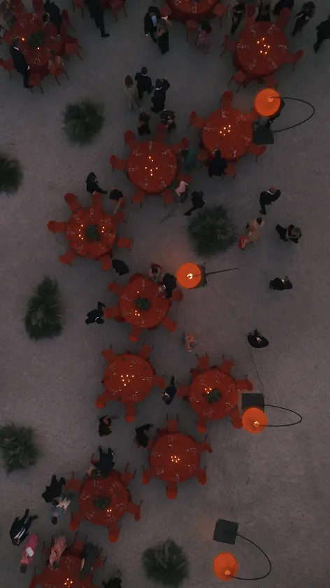 Aerial view of several round tables with orange chairs set outdoors on a stone surface, surrounded by people standing and walking, with illuminated orange street lamps and green bushes scattered around.