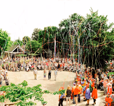 Large crowd gathered outdoors watching three people standing in the center as colorful streamers fall from above.