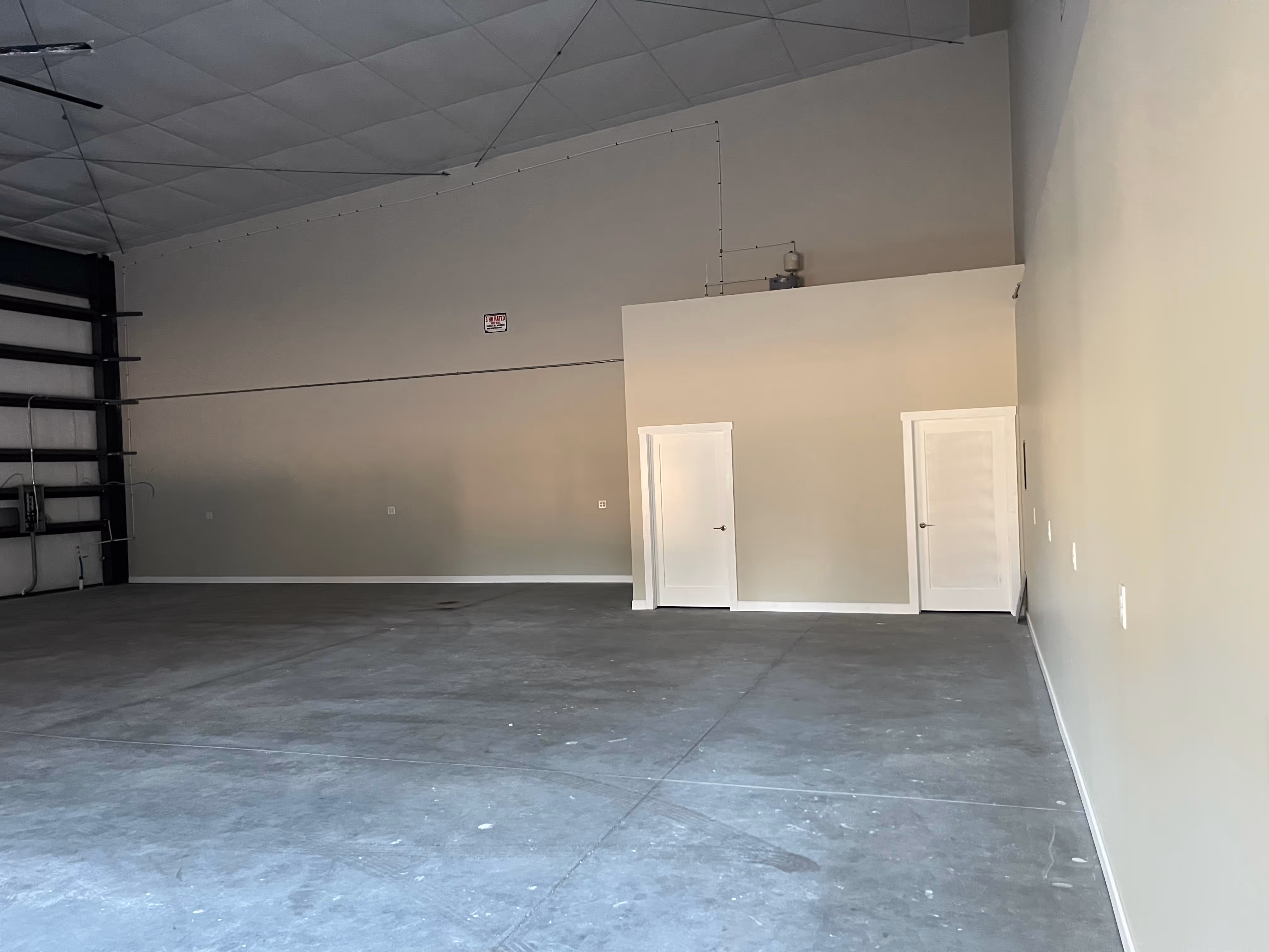 Empty industrial warehouse interior with concrete floor, high ceiling, and two white doors on a beige wall.
