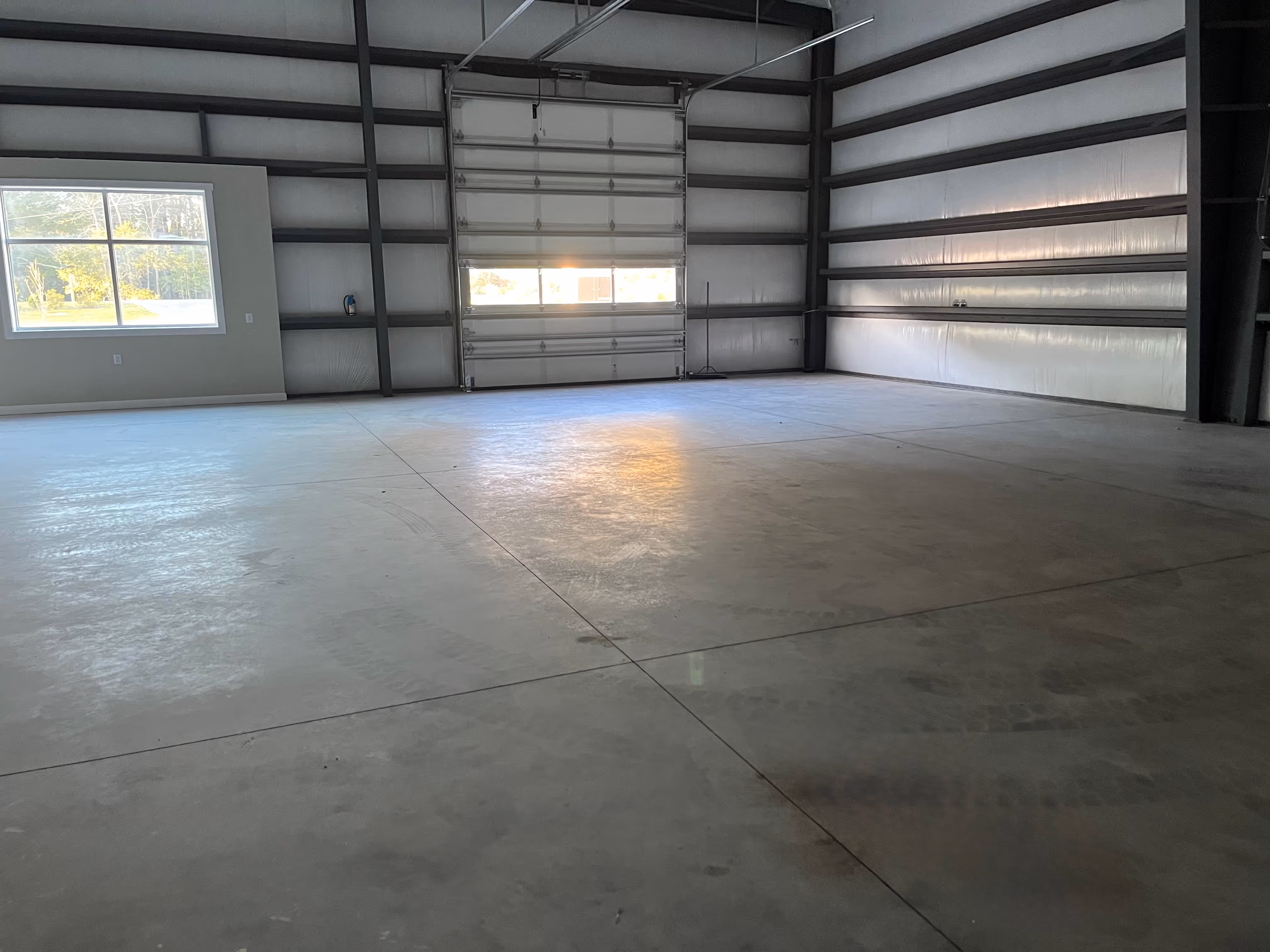 Empty industrial warehouse interior with concrete floor, metal walls, large overhead door, and a window showing greenery outside.