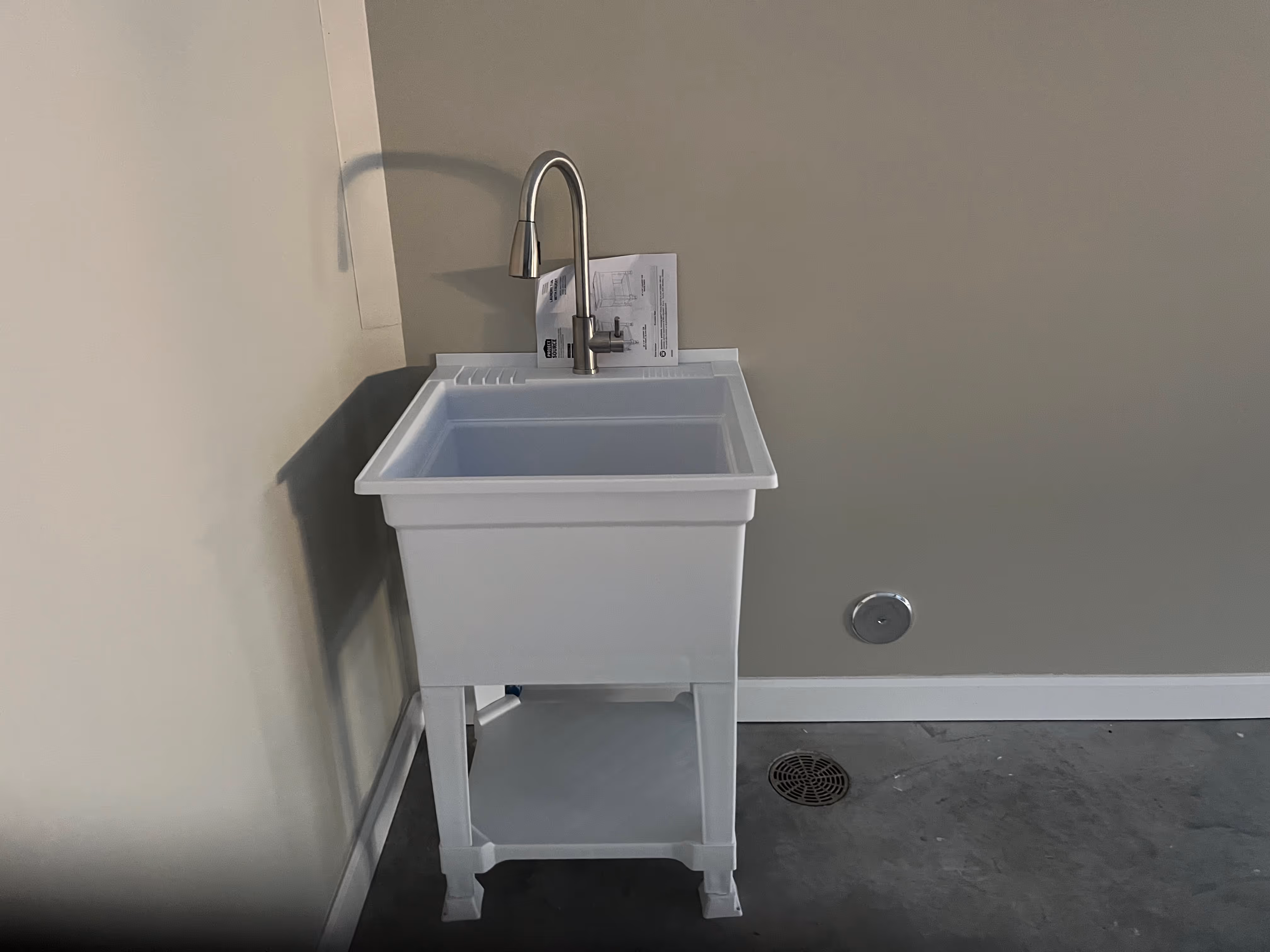 White utility sink with a stainless steel gooseneck faucet installed against a beige wall in a room with a concrete floor and a floor drain.