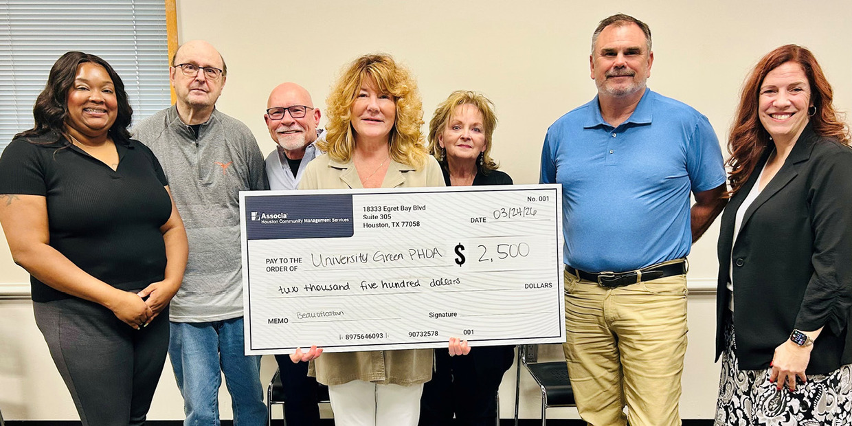 A group of six adults stand indoors in front of a plain wall, smiling at the camera while holding a large ceremonial check for $2,500 made out to “University Green PHCA.” The check is held across the center by three people, and the group is casually dressed.