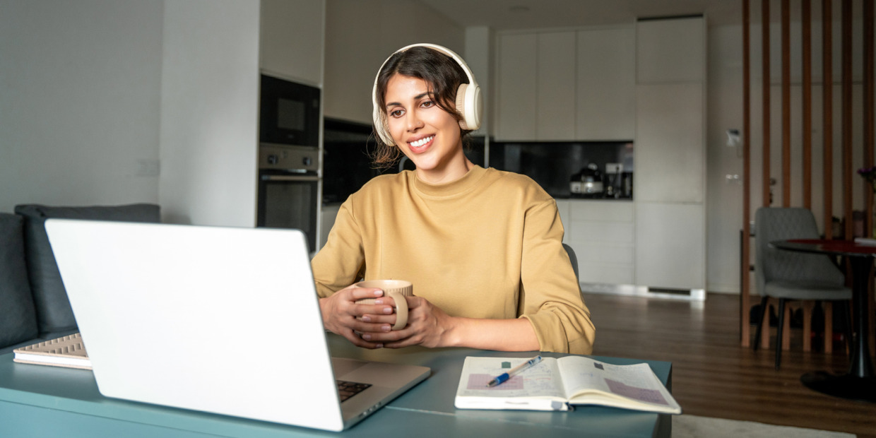 Woman wearing headphones smiles while participating in an online meeting on a laptop, holding a coffee mug at a desk in a modern home.