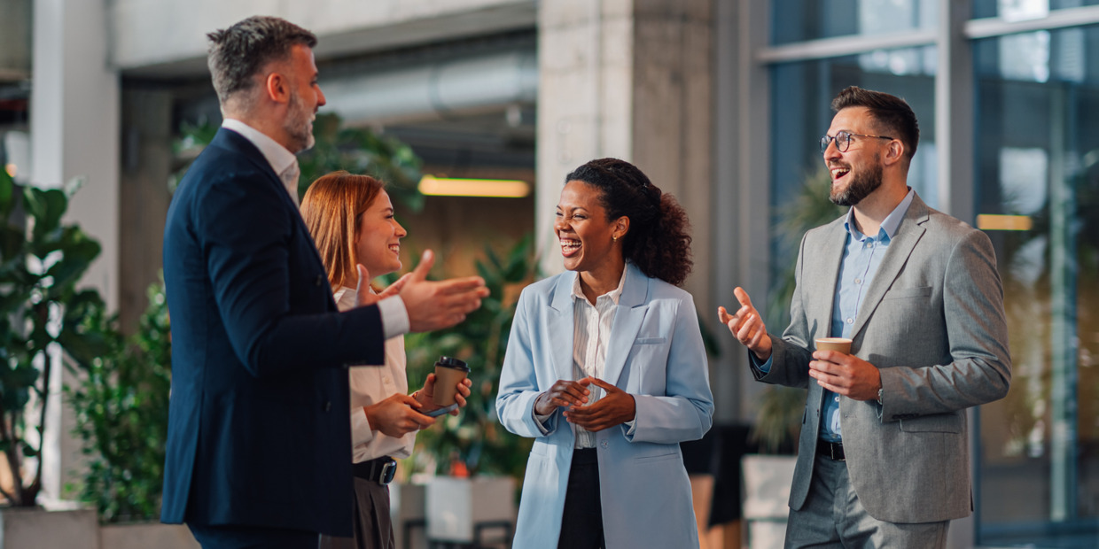 Four business professionals stand together in a modern office space, smiling and talking during a networking conversation. Two hold coffee cups while the group appears engaged in a friendly and collaborative discussion.