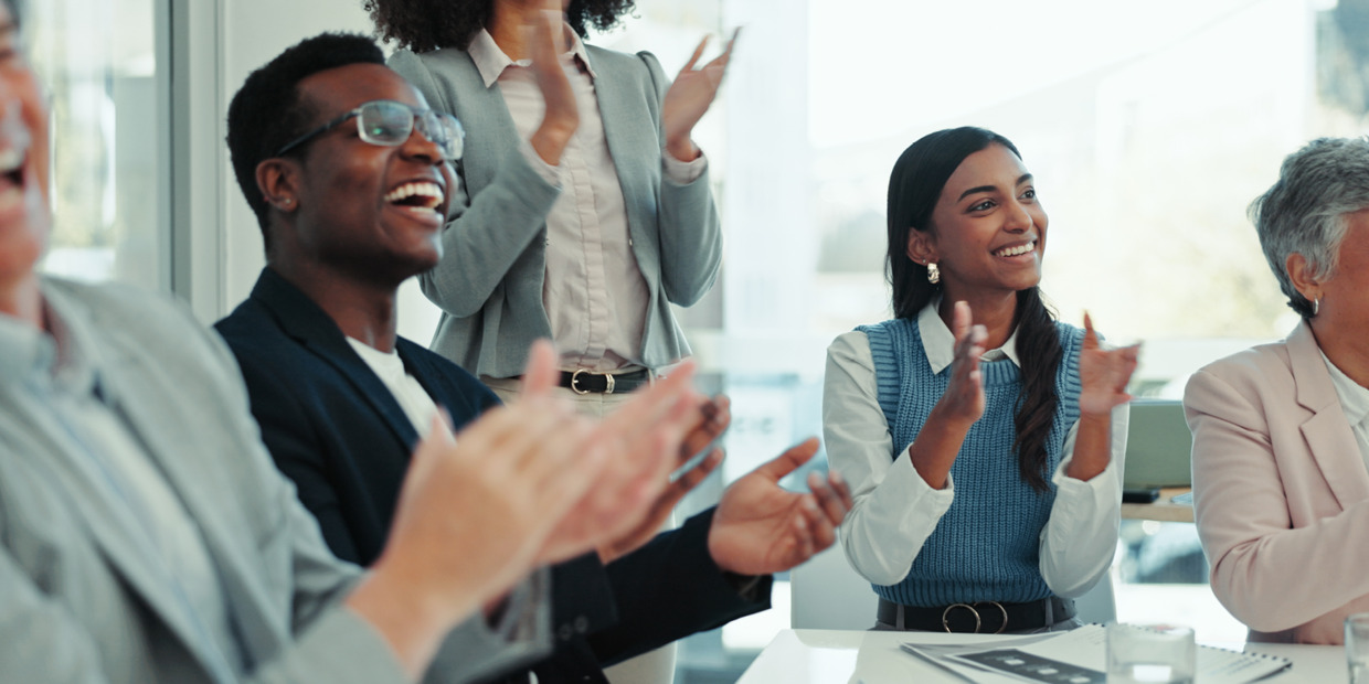 Diverse group of professionals seated and standing in a bright meeting space, smiling and applauding during a presentation or team event. The atmosphere appears positive and celebratory.