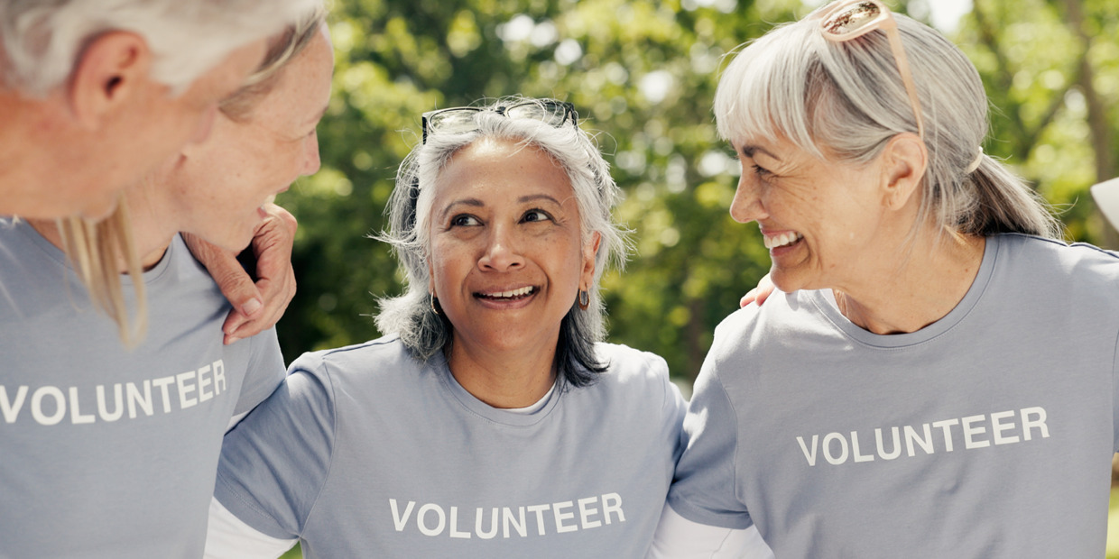 Three volunteers wearing gray shirts labeled “VOLUNTEER” smile and talk together outdoors.