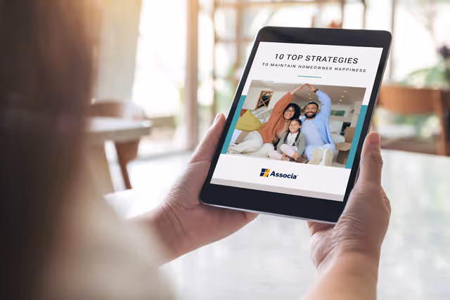 Person holding a tablet displaying an Associa eBook titled “10 Top Strategies to Maintain Homeowner Happiness” with a smiling family on the cover.

