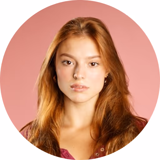 Portrait of a young woman with long red hair and a neutral expression against a pink background.