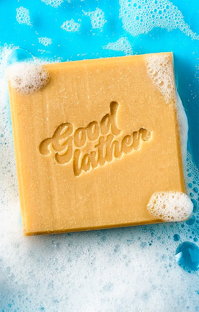 Vertical view of a square bar of Goodlather Lemongrass and Ginger CPG soap with a custom stamped logo floating in a bright blue pool of white suds, captured by Midnight Hour Studio.