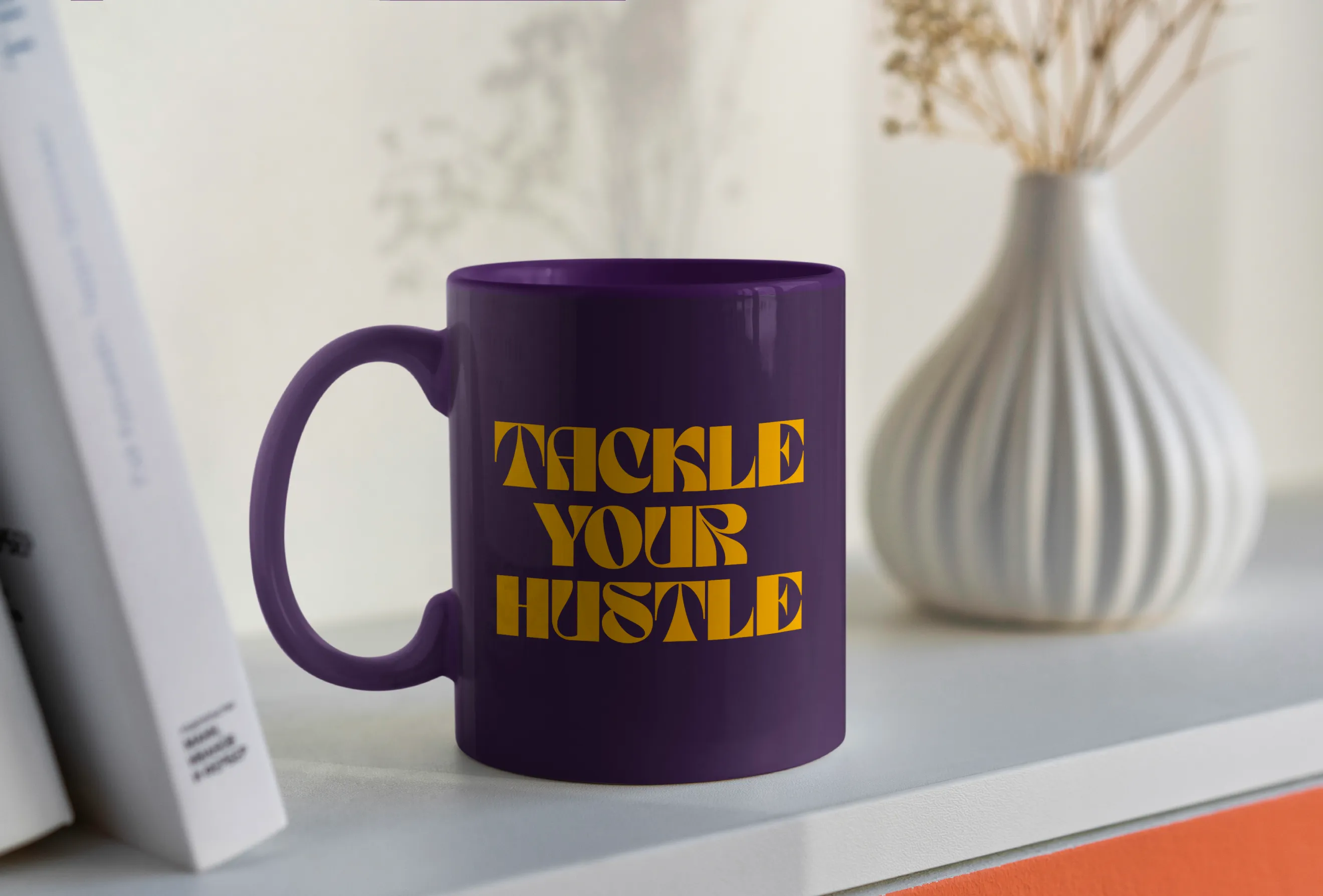 Deep purple ceramic mug on a white shelf beside books and a vase, featuring the bold orange phrase “TACKLE YOUR HUSTLE.”