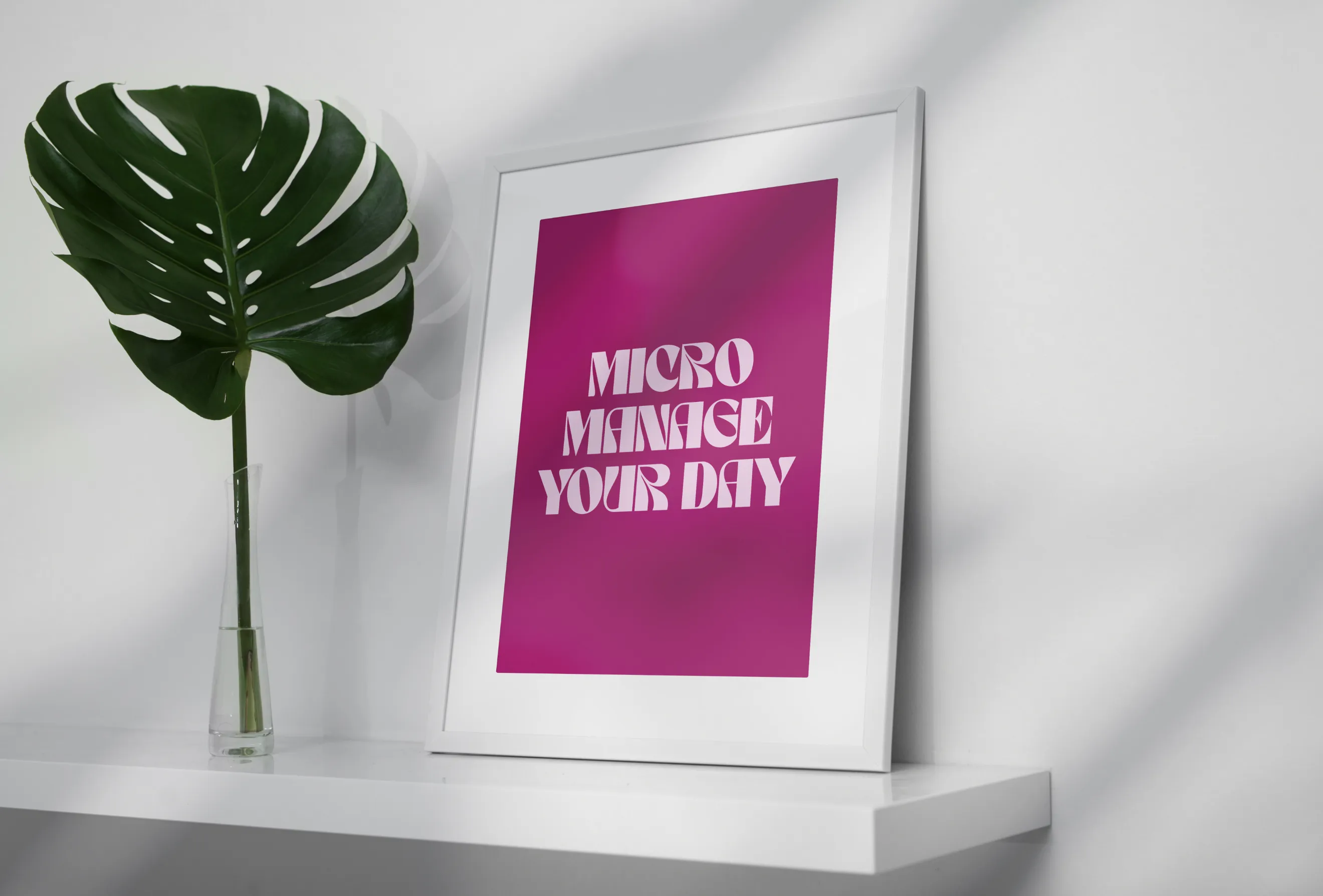 White-framed art print on a floating shelf beside a monstera leaf in a glass vase, featuring bold white typography on a rich magenta background.