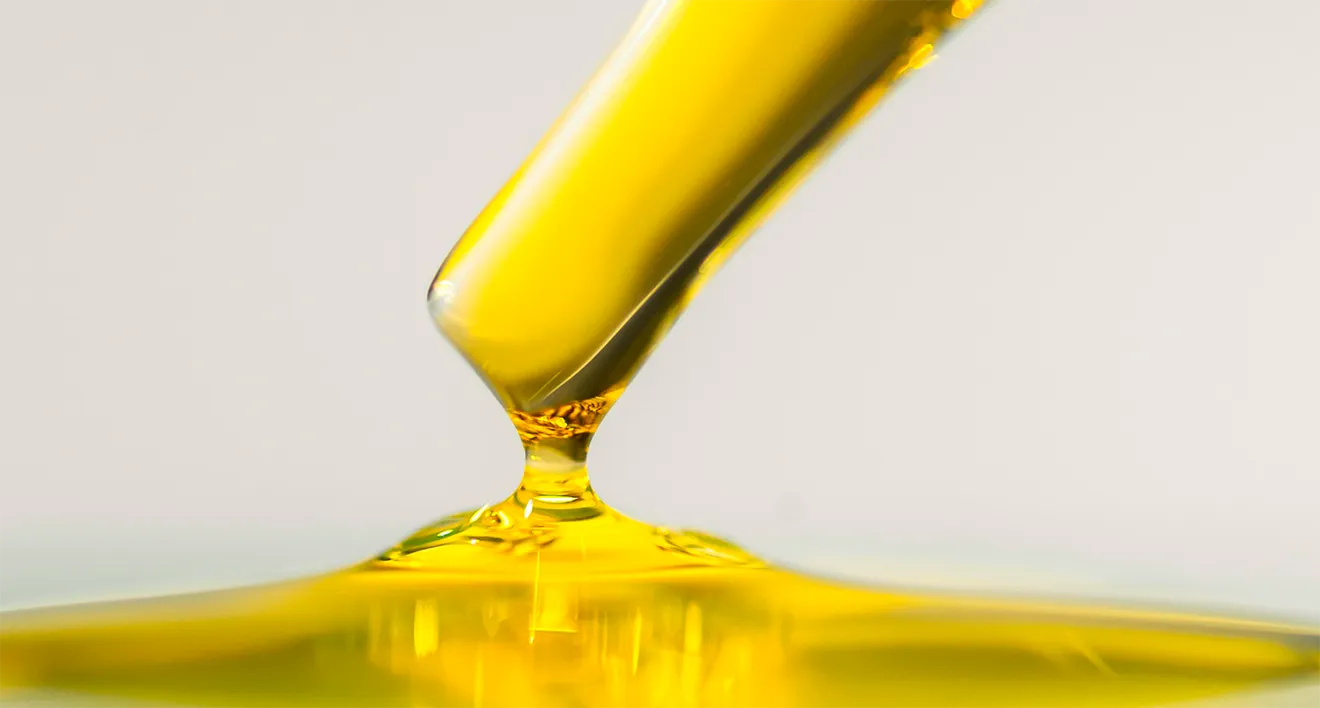 Macro photograph of a golden cannabis oil droplet on a glass pipette against a white background.