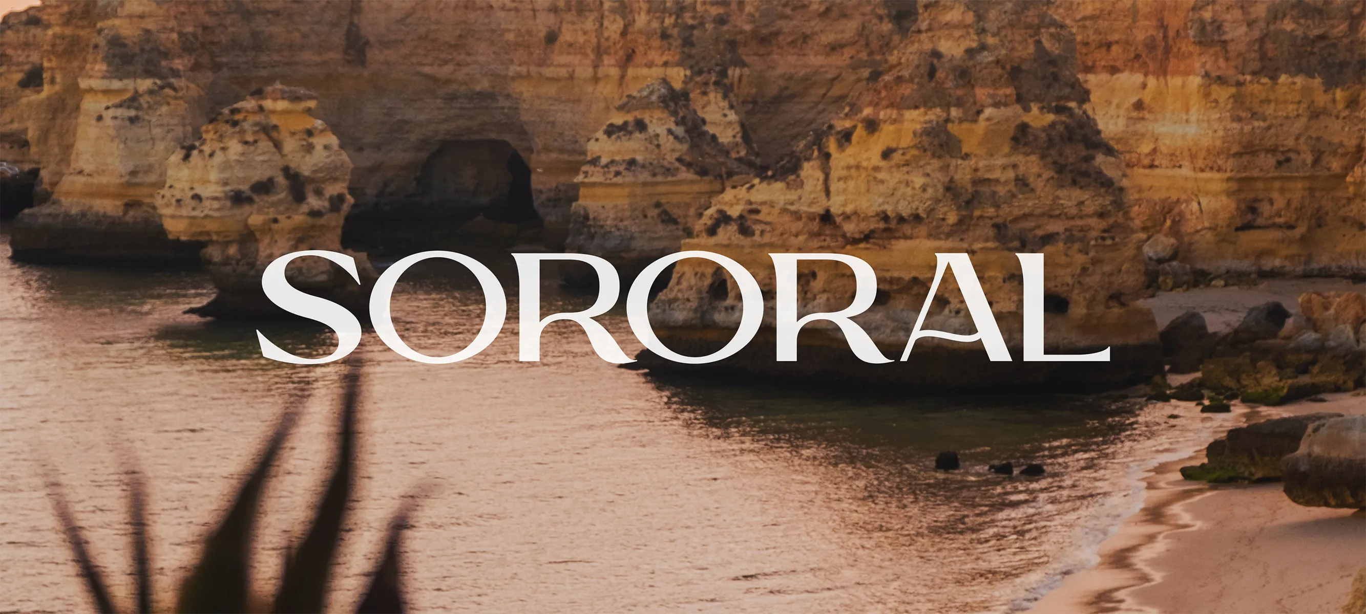 The word 'SORORAL' displayed in a bold, custom serif font with sweeping ligatures, overlaid across a scenic sunset view of rugged golden limestone coastal cliffs and sea caves meeting a calm beach.