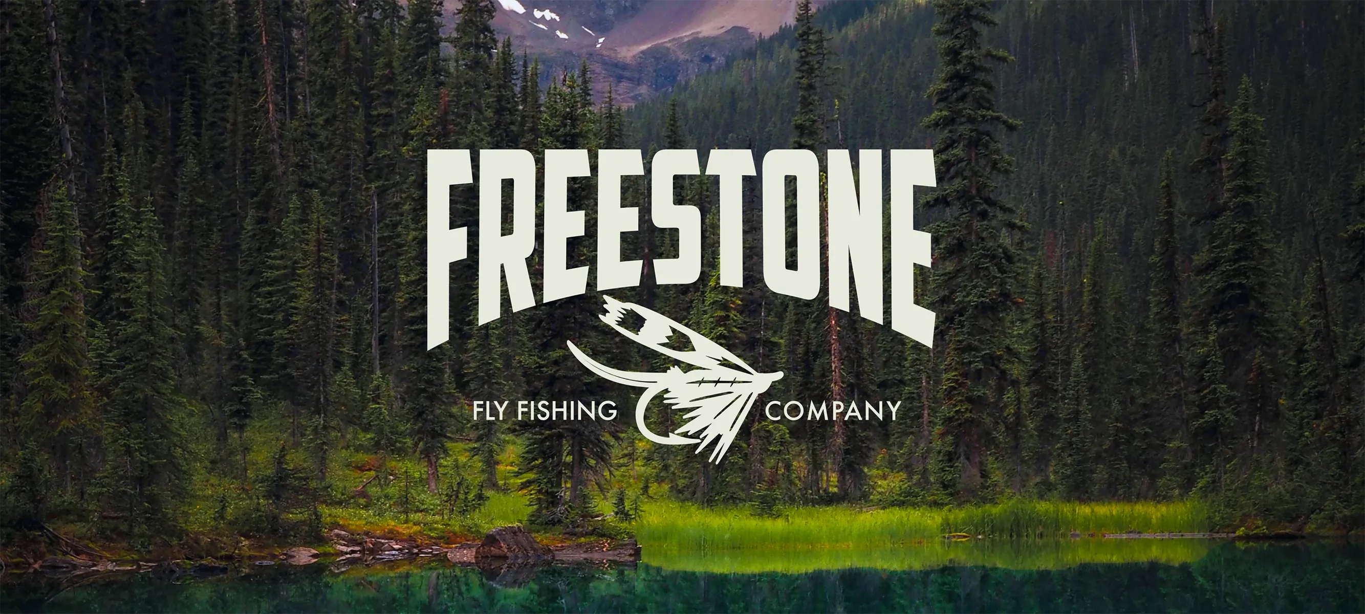 A wide website hero banner showcasing the white Freestone Fly Fishing Company logo lockup with a custom white hand-drawn fishing fly illustration. The graphic overlay is centered over a panoramic landscape of a dense, moody evergreen forest reflecting into a calm Canadian lake with mountains in the background.
