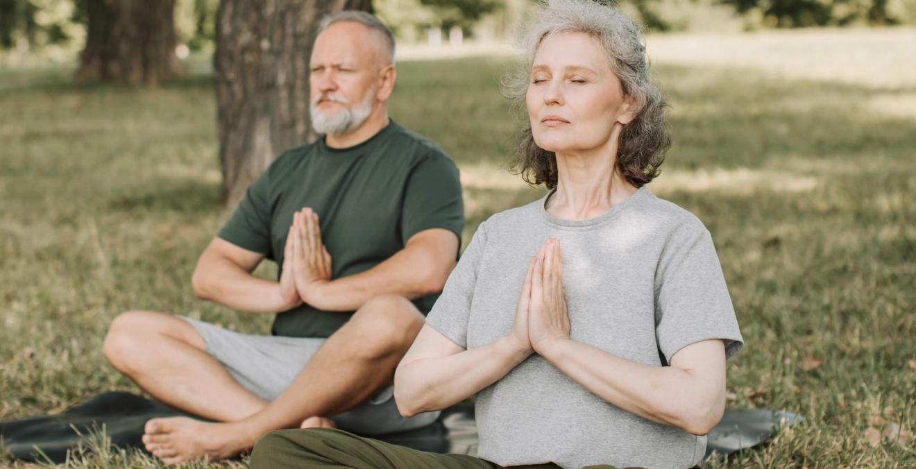 Older couple meditating outdoors on yoga mats, representing inclusive wellness and mindful self-care.
