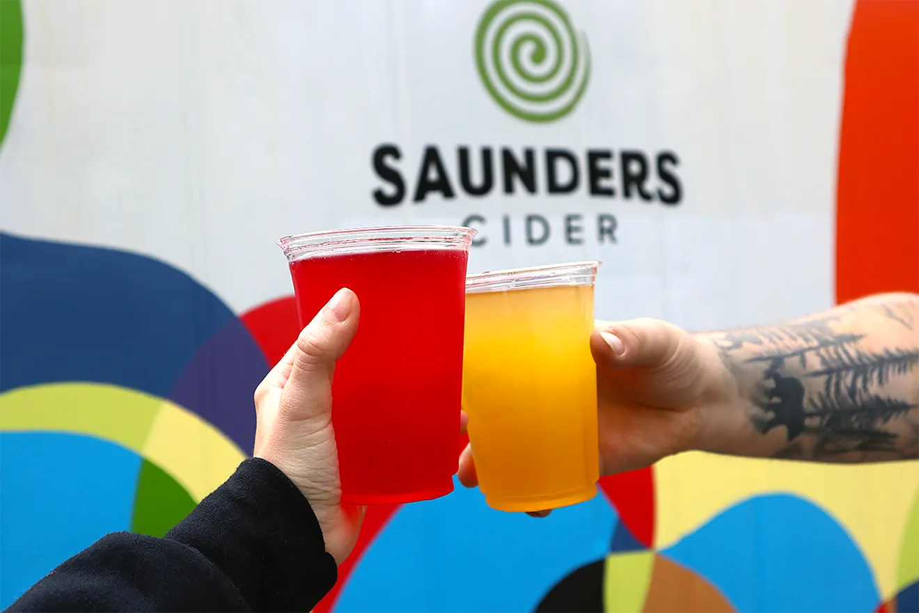 First-person photo of two hands toasting with Saunders Cider cups in front of an event backdrop by Hovski Studio.