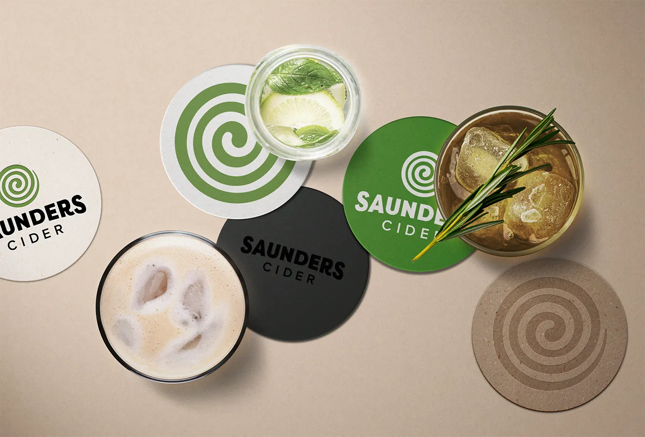 Overhead flat lay of five custom Saunders Cider drink coasters by Hovski Studio alongside iced cider glasses.