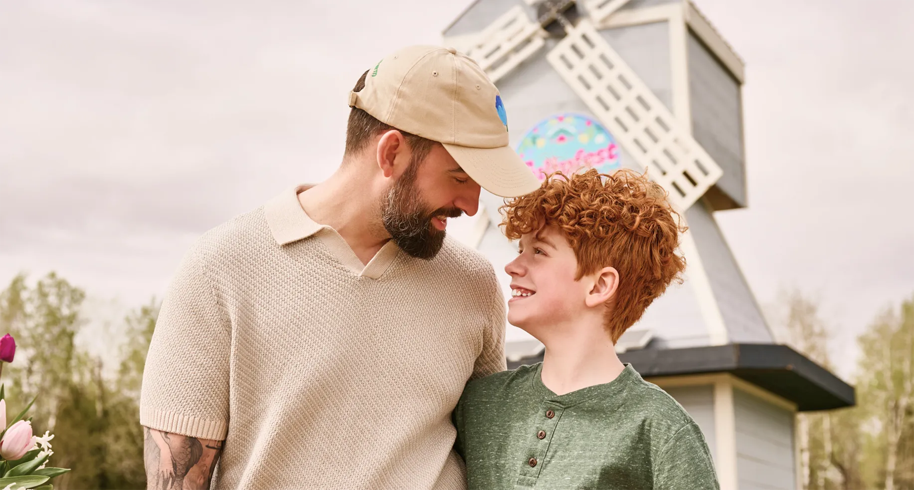 A man wearing a tan Tulipfest embroidered cap looks down smiling at his curly-haired son in front of a large, custom-built grey and cream windmill. A circular, colorful Tulipfest brand sign is mounted directly onto the front facade of the windmill structure, signage designed by Hovski Studio.
