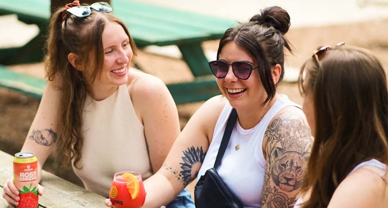 Three women laughing together at outdoor picnic table enjoying Saunders Cider Rosy Cheeks craft cider.
