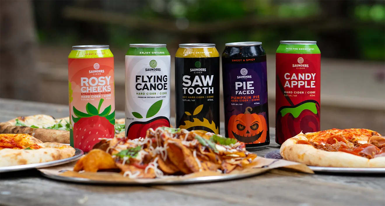 Lineup of Saunders Cider craft beverage cans including Rosy Cheeks, Flying Canoe, Saw Tooth, Pie Faced, and Candy Apple on patio table.
