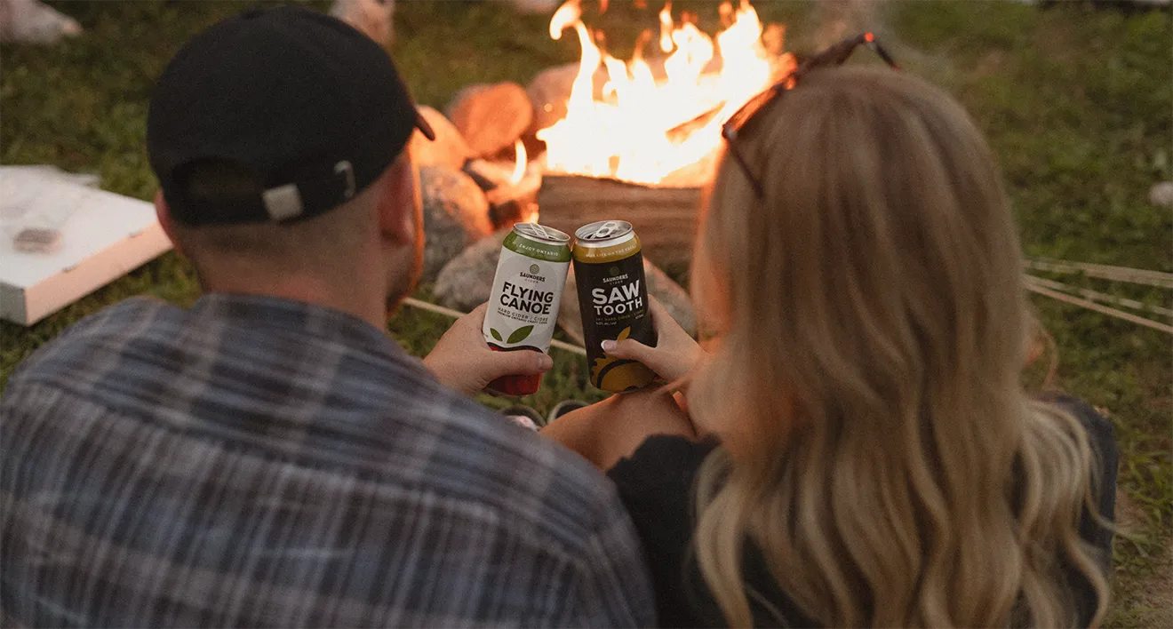 Couple enjoying Saunders Cider Flying Canoe and Saw Tooth craft cider cans around a outdoor campfire.