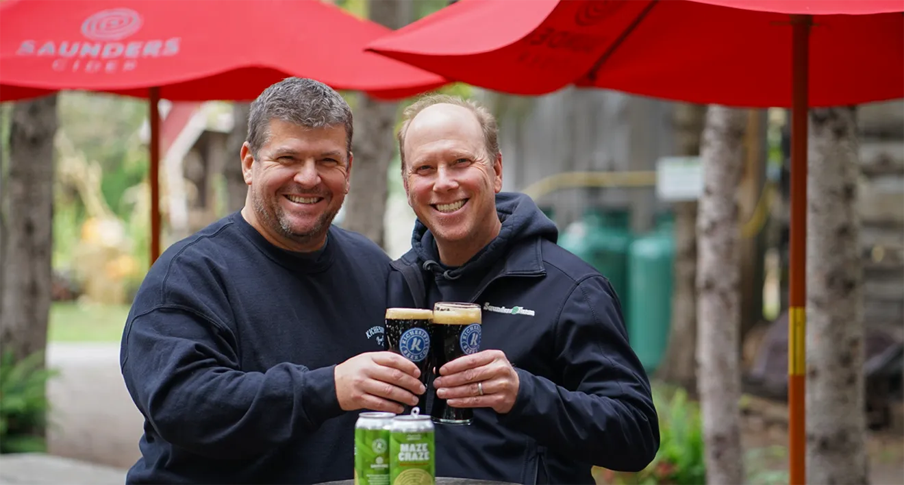 Saunders Farm owner and Paul Meek toasting with craft beer and cider glasses on outdoor patio under red umbrellas.