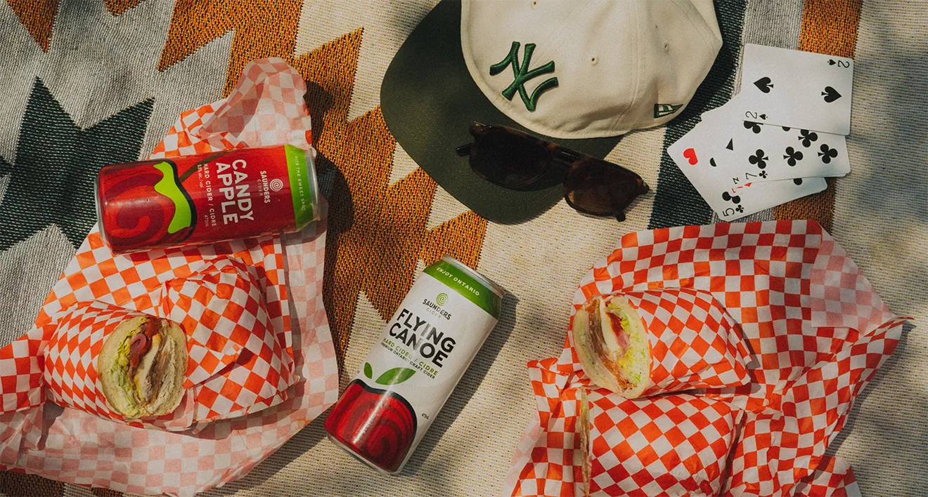 Retro outdoor picnic flatlay with Saunders Cider cans, wrapped sandwiches, playing cards, and sunglasses.