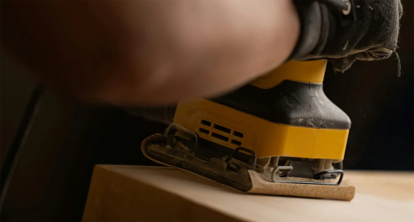 Woodworker using electric orbital sander on custom raw wood furniture piece.